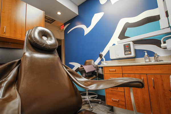 An image of a modern dental office interior featuring a brown dental chair with a large, circular headrest and a blue wall with a white and black abstract design.