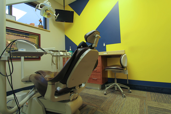 The image shows an interior view of a dental office, featuring a dental chair with a patient s headrest and a dental machine in the foreground. The room has a blue wall with a yellow abstract design on one side, a reception desk with a computer monitor and keyboard, and a sign that reads  Dental Office  above the desk. There is a window with blinds partially drawn.