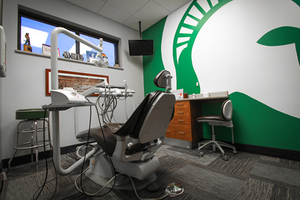 The image depicts a dental office interior with a wall mural featuring the university s logo and a large, modern dental chair.