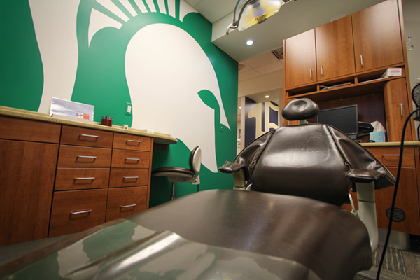 The image shows an interior dental or medical office with a prominent Spartan logo on the wall, a dental chair in the foreground, and various pieces of dental equipment.