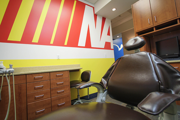 The image depicts a dental office interior, featuring a dental chair with an adjustable headrest and a dental mirror in front of it.