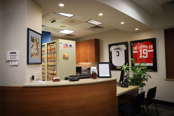 The image depicts an interior view of a well-lit office space, featuring a reception area with a desk and chairs, framed pictures on the wall, and a large window allowing natural light to enter.