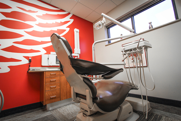 The image shows an interior view of a dental office, featuring a modern dental chair with adjustable headrests and a red wall with a decorative pattern in the background.