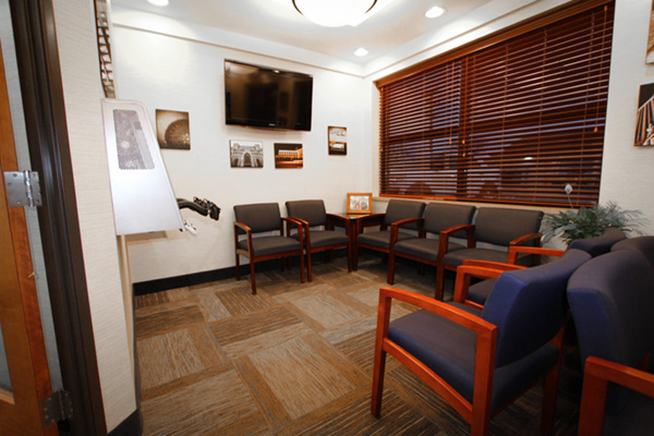 The image depicts an office waiting room with a modern design, featuring a carpeted floor, wooden chairs, a television mounted on the wall, and framed pictures or certificates.