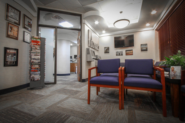 The image shows an interior space, likely a waiting room, with a carpeted floor, a reception desk, chairs, and a framed certificate on the wall.