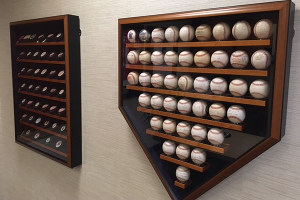 The image shows a wall-mounted display case containing numerous baseballs and a small collection of trophies, with the balls arranged in rows and the trophies placed at the top.