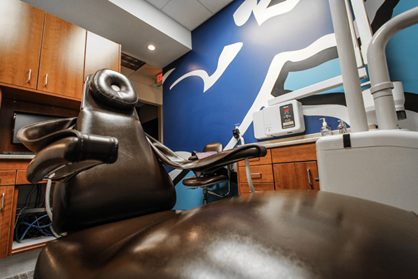 The image shows a dental office interior with a chair, a sink, and a wall-mounted television displaying an abstract design.