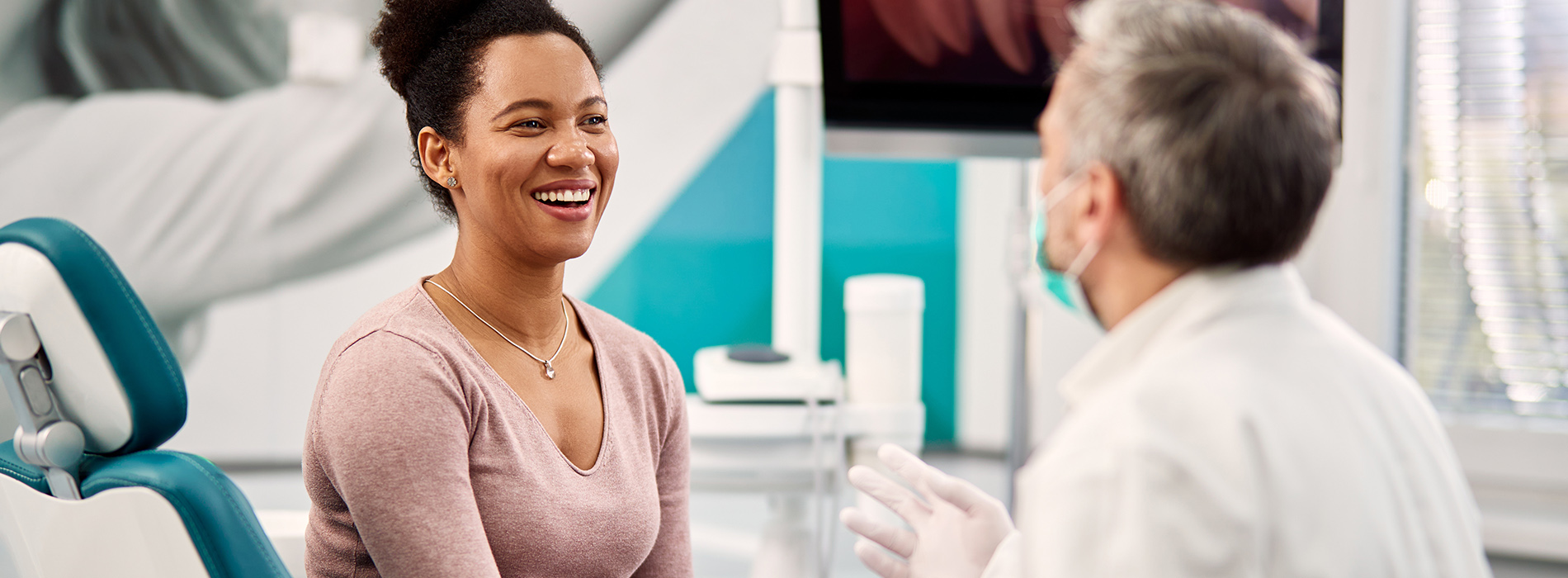 A woman in a dental office, smiling and seated in front of a dentist.