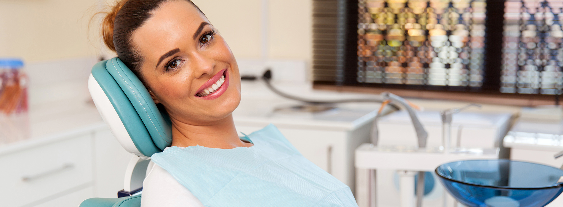 A woman sitting in a dental chair with a smile, wearing a blue surgical gown and a white headset.