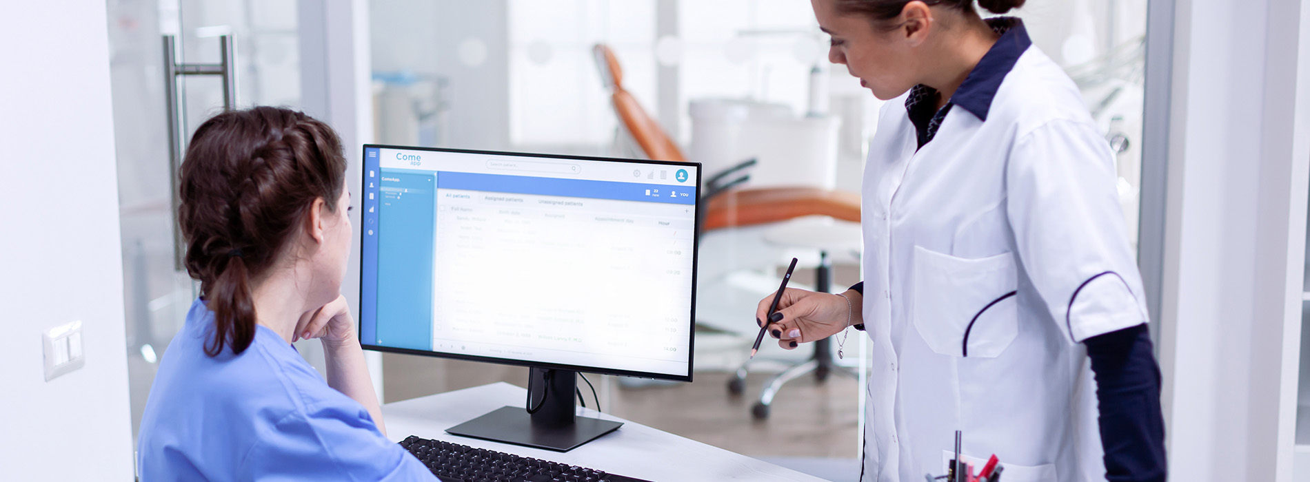 The image displays a professional setting with a healthcare worker standing behind a desk, interacting with a computer screen, while a patient sits on the other side of the desk. The environment suggests a modern medical office or clinic.