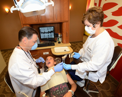 An image of a dental office setting where a dentist is performing a procedure on a patient while being assisted by a hygienist.