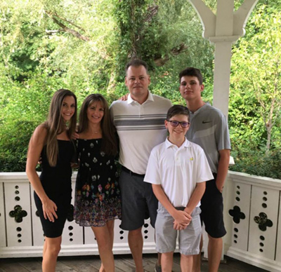 The image depicts a family of four, including an adult male, two females, and a young boy, posing together for a photo on what appears to be a balcony or porch with a scenic backdrop.