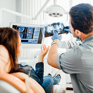 A dental professional is showing a patient an X-ray of their teeth, using a large monitor in a dental office setting.