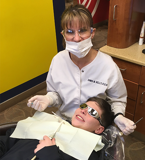 An adult dental patient receiving care while a dental hygienist assists, both wearing protective eyewear and following standard dental office safety protocols.