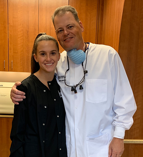 In the image, a man and a woman are posing for a photo together. The man is wearing a white lab coat and has a stethoscope around his neck, while the woman is smiling at the camera. They appear to be in an office or medical setting, as suggested by the presence of a desk with a computer monitor and other items in the background.
