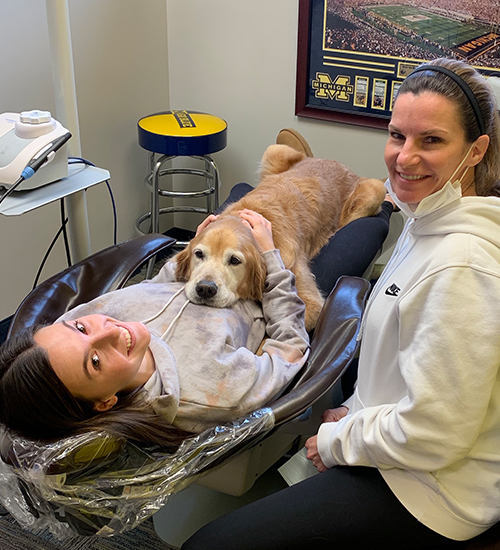A woman and a dog are sitting in dental chairs, with the woman holding the dog s head.