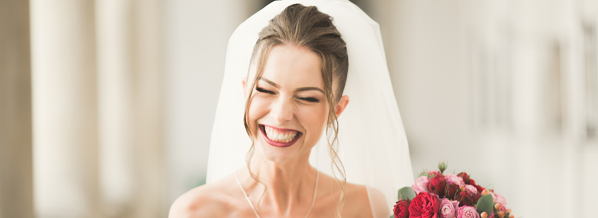 A woman in a white wedding dress smiling, with a veil and holding a bouquet.