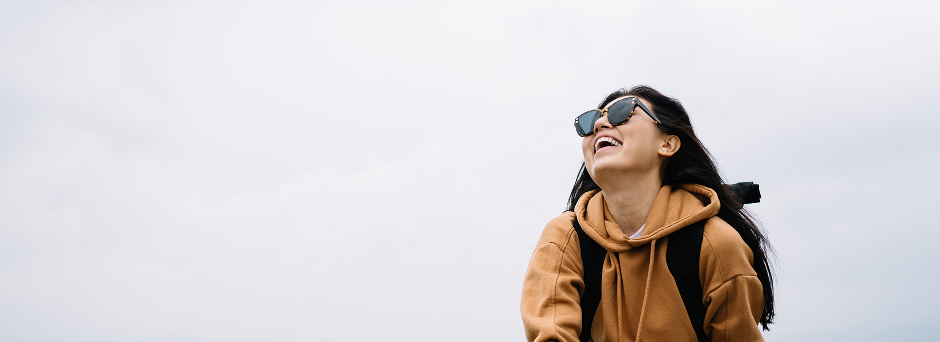 A woman in a hoodie and sunglasses, smiling at the camera against a cloudy sky background.