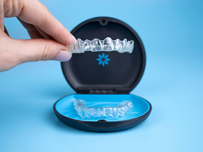 A person s hand is holding a clear plastic tray with a blue lid, revealing a set of transparent dental trays inside.