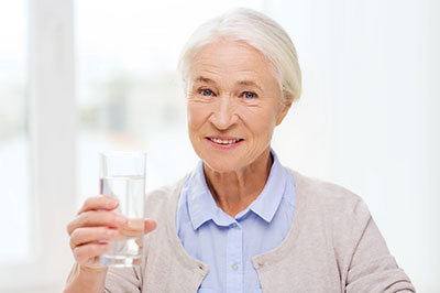 An elderly woman in a blue blouse holding a clear glass of water while smiling.