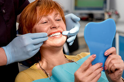 A woman in a dental chair is smiling while looking into a mirror, with a dental hygienist adjusting her teeth.