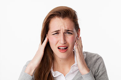 The image shows a woman with her hand on her head, holding her ear as if she is experiencing discomfort or pain, with an expression of concern or distress.