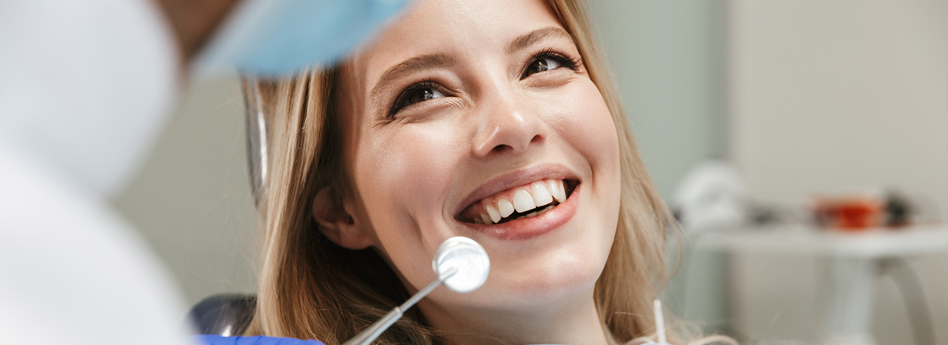 A smiling woman in a dental chair, looking at the camera, with a dentist in the background.