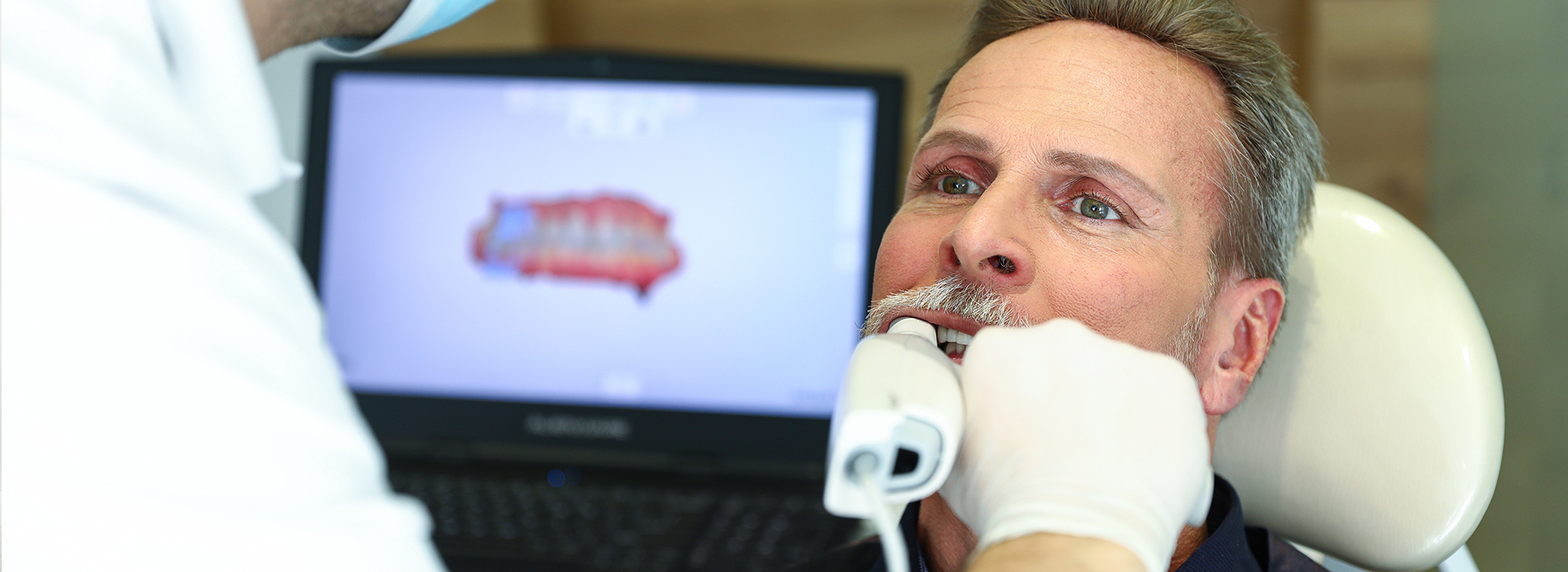 The image shows a man receiving dental treatment, with a dental professional using a computer screen to guide the procedure.