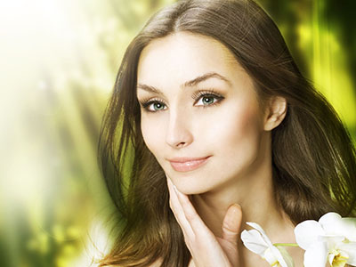 The image features a woman with fair skin, wearing makeup that includes lipstick and eye shadow. She has long brown hair and is looking directly at the camera. Her expression appears calm and serene. The background is blurred but suggests an outdoor setting with natural elements like foliage and possibly a water feature or fountain.