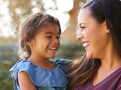 A woman and a young girl are smiling, with the child holding onto the woman s leg. They appear to be outdoors during daylight.