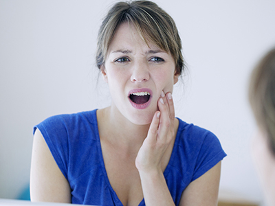 The image shows a woman with her mouth open, expressing surprise or shock, while holding her hand to her face near her nose. She is looking at her reflection in the mirror.