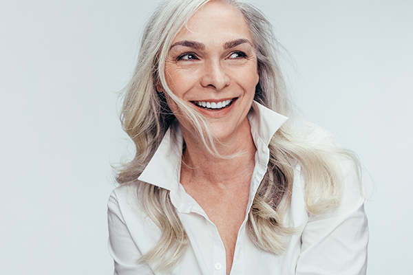 The image shows a woman with short blonde hair, wearing a white top with the sleeves rolled up to her elbows. She has a neutral expression on her face and is looking directly at the camera. Her posture is relaxed with one hand casually resting on her hip.