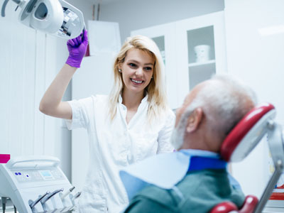 Woman in white uniform assisting elderly man with dental equipment.