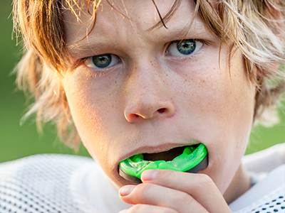 A young boy with blonde hair and a determined expression is holding a green toothbrush to his mouth, possibly in the midst of brushing his teeth.