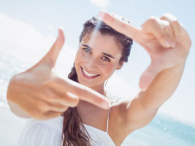 A woman with a radiant smile is holding up her hand, framing the camera lens, against a bright background.