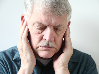 The image shows an elderly man with a mustache, holding his hand to his ear as if experiencing discomfort or pain.